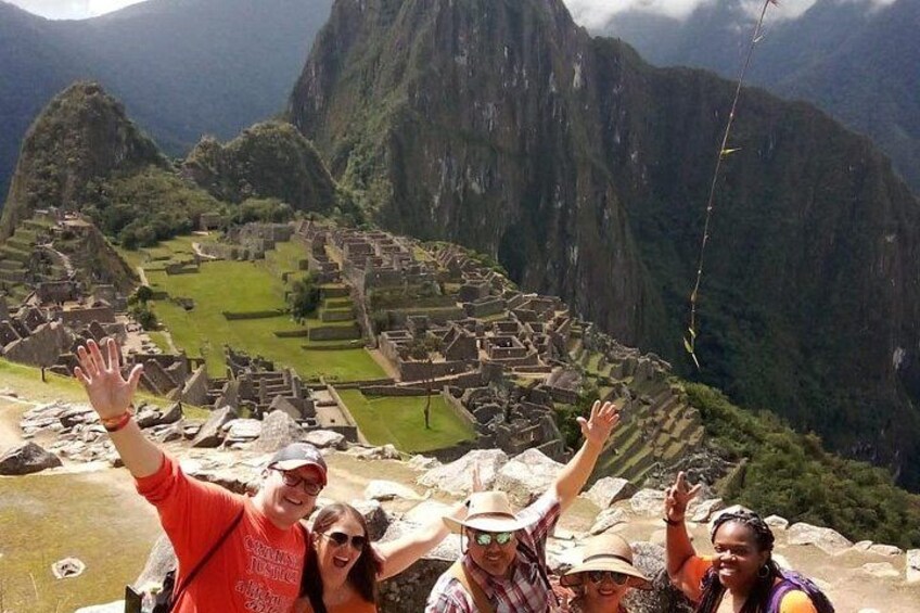 A group of people standing on a rocky outcrop with their arms raised, overlooking the ruins of Machu Picchu. 3