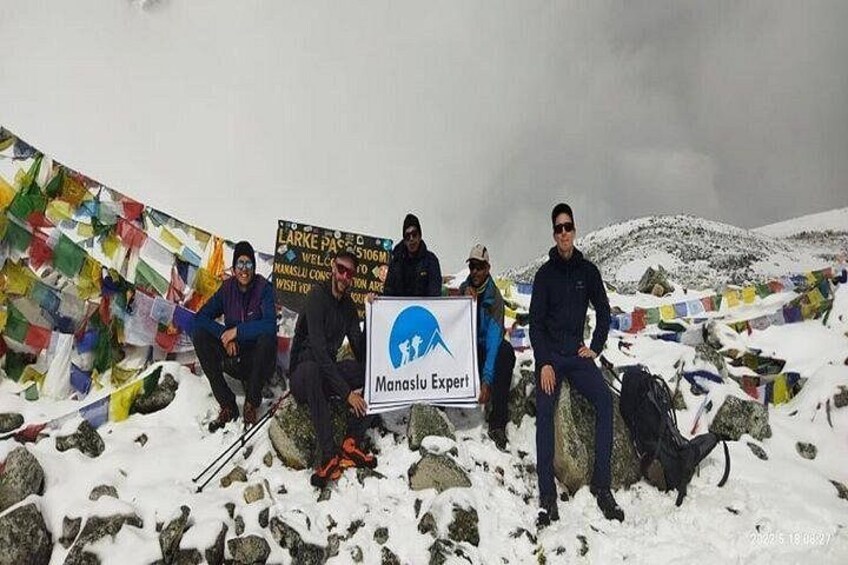 En grupp människor på ett snötäckt berg, med böneflaggor och en skylt med texten ”Manaslu-expert”. 2