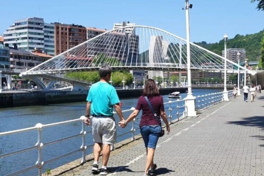 A couple walking on a bridge with a river, a cityscape with buildings, and a clear sky. 3