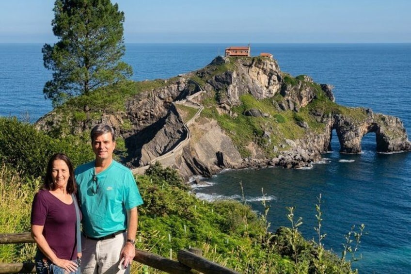 A coastal landscape with a rocky outcrop, a sea arch, and a small building on top. 1