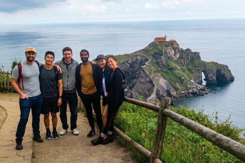A group of people standing on a path overlooking a coastal landscape with a prominent cliff and a small building on top. 5