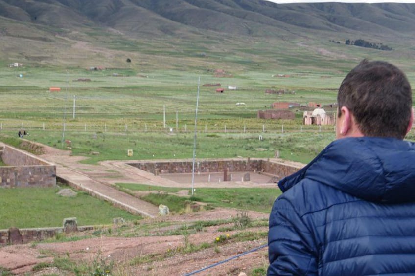 Un hombre con una chaqueta azul observa un paisaje rural con campos, una estructura de piedra y edificios al fondo. 2