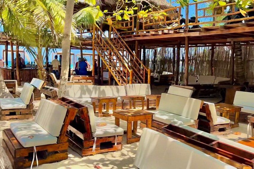 A beachside lounge with wooden furniture, palm trees, and a staircase leading to an upper deck. 4