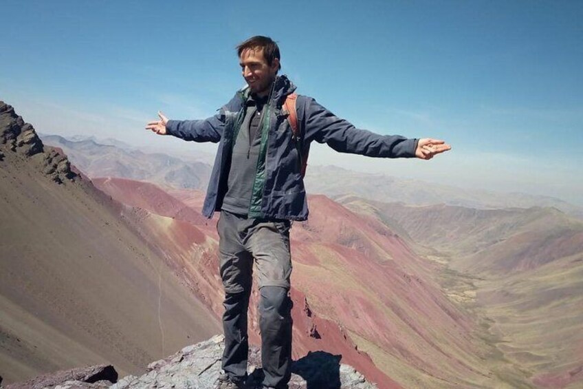 Una persona de pie en la cima de una montaña con los brazos extendidos, vestida con una chaqueta y pantalones, con un cielo despejado y un paisaje montañoso al fondo. 4