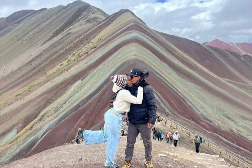 Una pareja se abraza en la cima de una montaña, con formaciones rocosas de colores al fondo. 2
