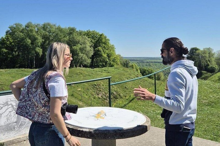 Dos personas de pie junto a una mesa, con un paisaje al fondo. 1