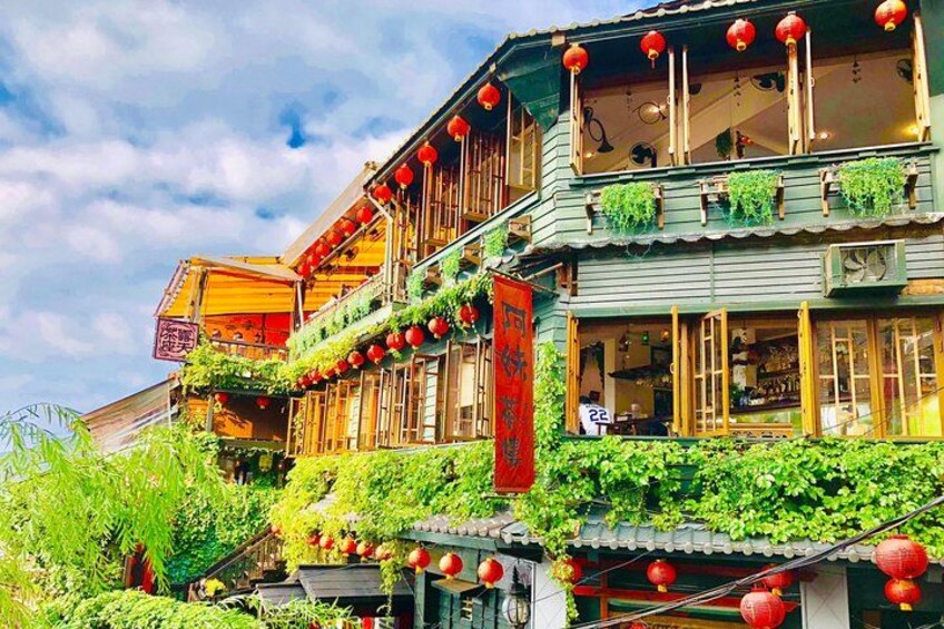 A traditional building with green walls, red lanterns, and plants growing on the exterior. 1