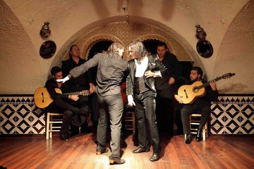 A group of people in a room with wooden flooring and patterned walls, some holding guitars. 2