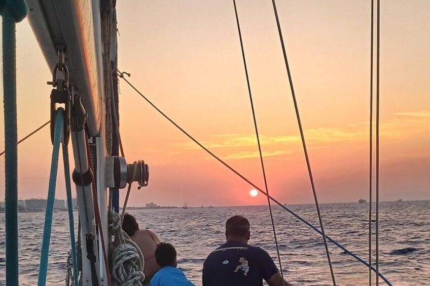Atardecer sobre el océano con un velero, dos personas en la cubierta y un horizonte urbano a lo lejos. 4