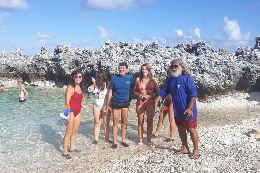 A group of people standing on a pebbly beach with clear water and rocky formations in the background. 2
