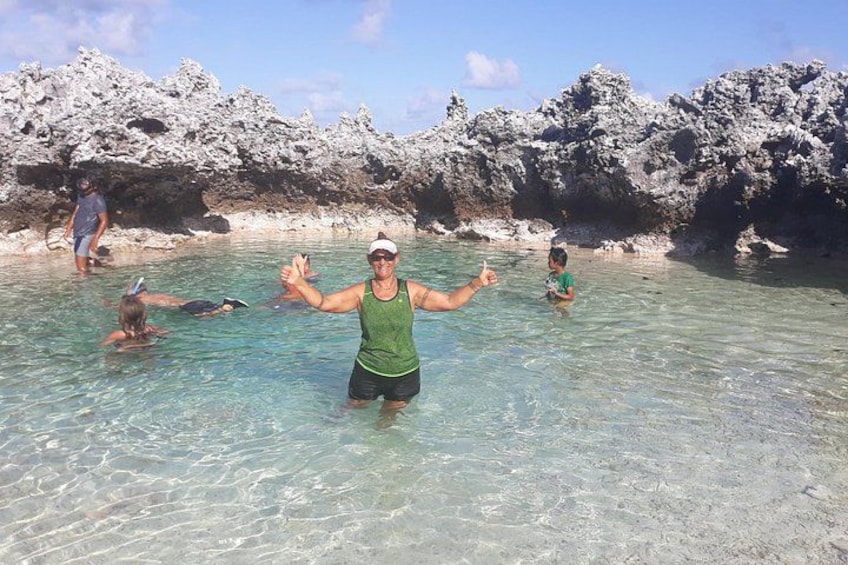 Une personne portant un débardeur vert et un short noir, debout dans une eau peu profonde, avec une côte rocheuse en arrière-plan. 2
