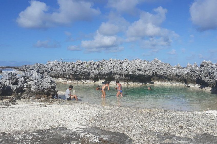 Un rivage rocheux bordé d’une eau claire, où l’on voit des gens nager et une personne assise sur le sable. 3