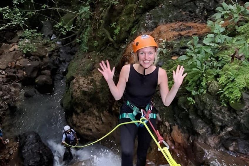 A person rappelling down a rocky cliff with a harness and helmet, surrounded by greenery and a stream. 1