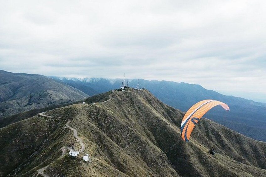 A mountainous landscape with a person paragliding, a trail, and distant snow-capped peaks. 4