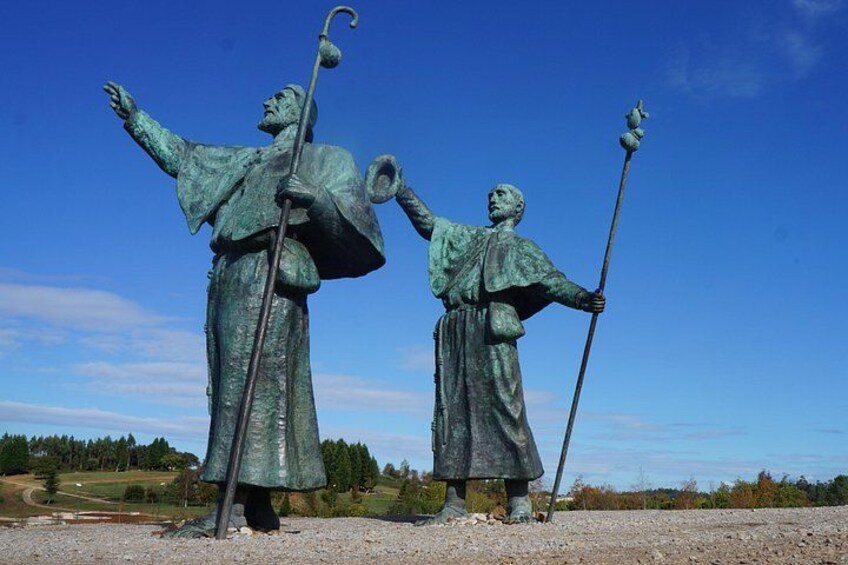A statue of two figures with outstretched arms and staffs, set against a clear blue sky and a rural landscape. 3