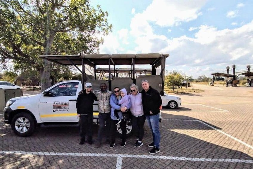 A group of people standing in front of a white van with a canopy, parked on a paved area with trees and a clear sky in the background. 4