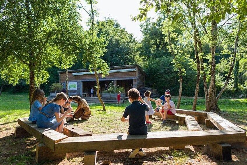 A group of people sitting on benches in a park with trees and a wooden structure in the background. 5