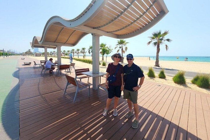 A wooden boardwalk with a curved shelter, palm trees, and a beach in the background. 2