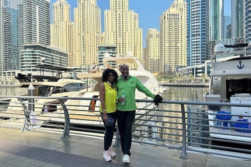 A marina with yachts, a city skyline, and a person in a green jacket leaning on a railing. 4