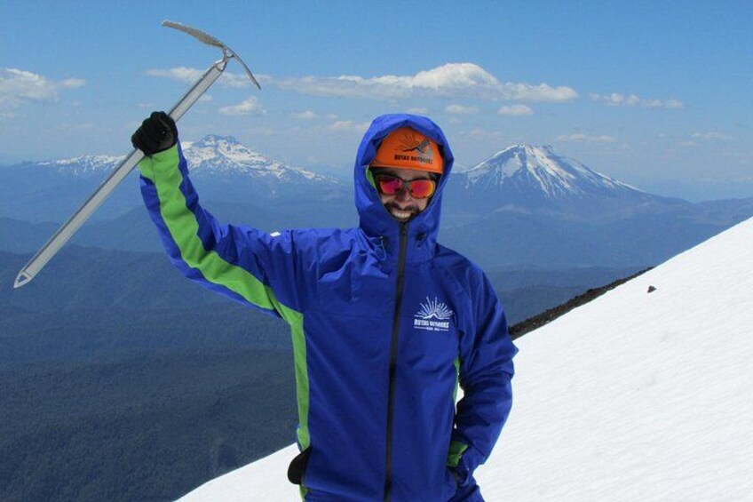 Una persona con una chaqueta azul sostiene un bastón de esquí, de pie en una ladera nevada con una montaña al fondo. 1