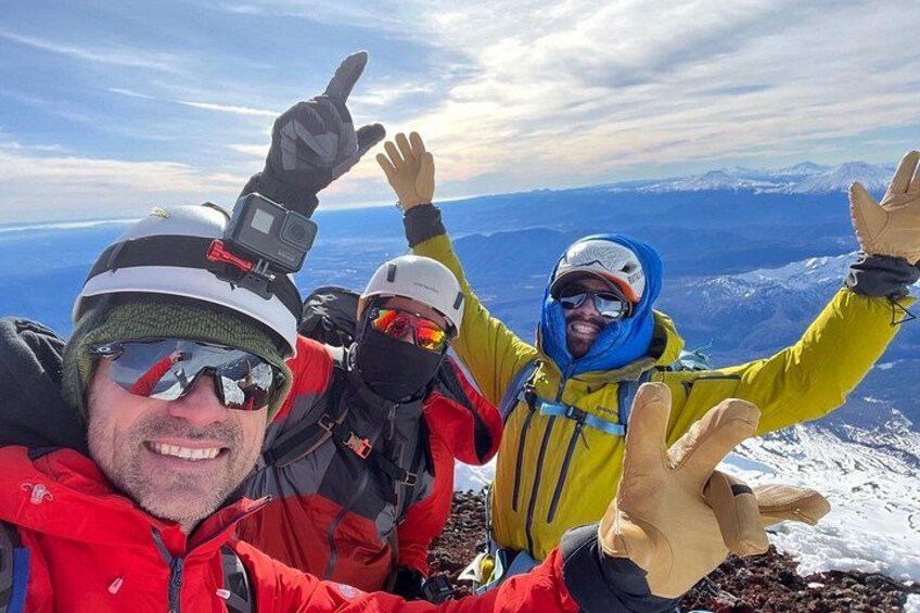 Tres personas en la cima de una montaña, con cascos y chaquetas, con un cielo despejado y un paisaje nevado al fondo. 5