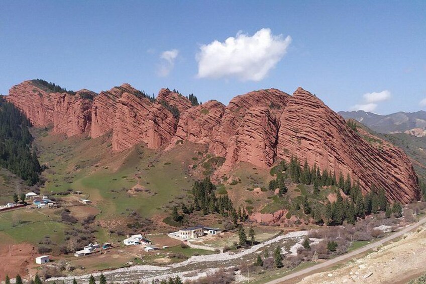 A mountainous landscape with red rock formations, a small settlement with buildings, and a clear sky. 3