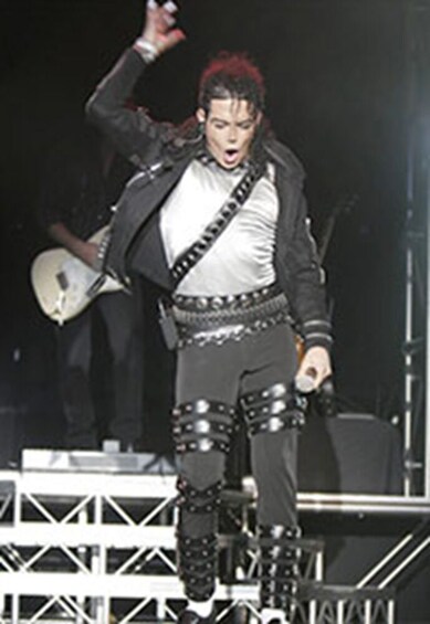 A person on stage with a guitar, wearing a black jacket, white shirt, and black pants with buckles. 5