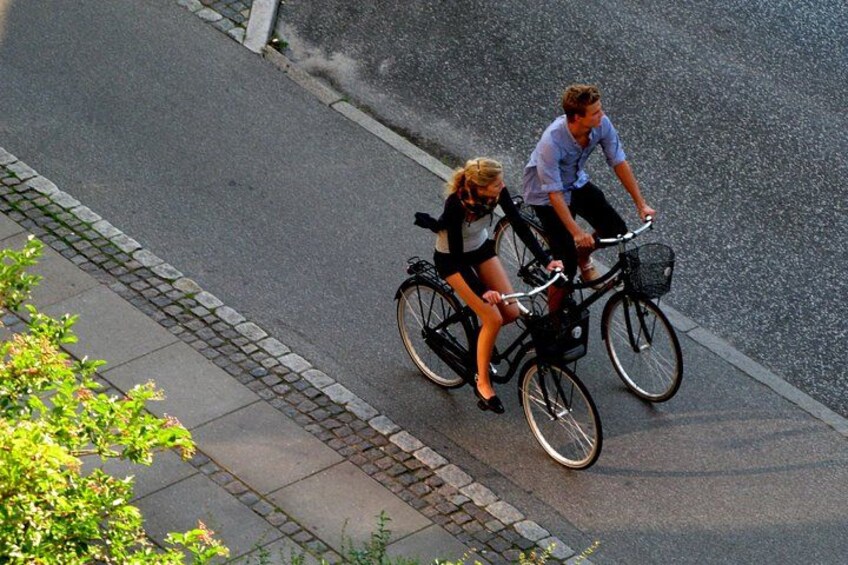 Two people riding bicycles on a paved road with a curb and a sidewalk. 5