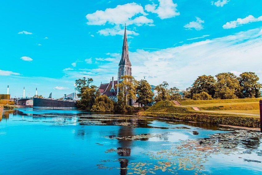 A church with a tall spire, a river, and a park with trees. 1