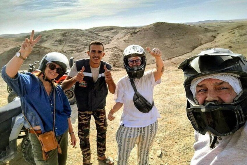Four people in desert attire with helmets, standing on a dirt road with a vehicle in the background. 5