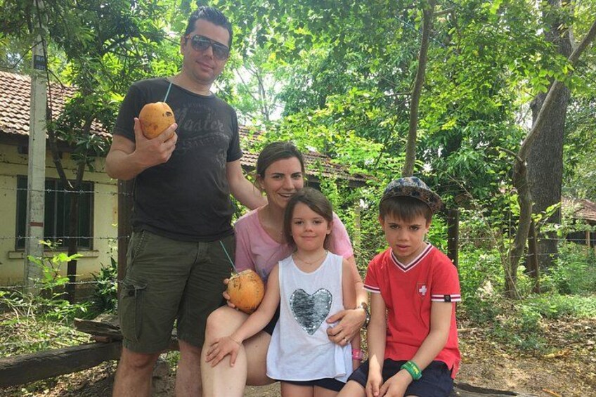 Une famille de quatre personnes en plein air : un homme tient un fruit, une femme porte un chemisier rose, une fille porte un chemisier blanc et un garçon porte un t-shirt rouge. 1