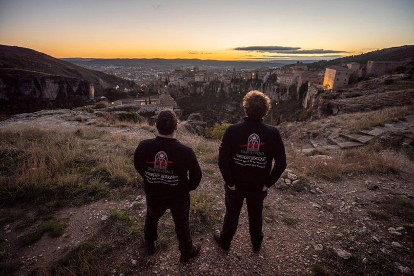 Two people standing on a rocky path overlooking a valley with buildings and a sunset in the background. 5