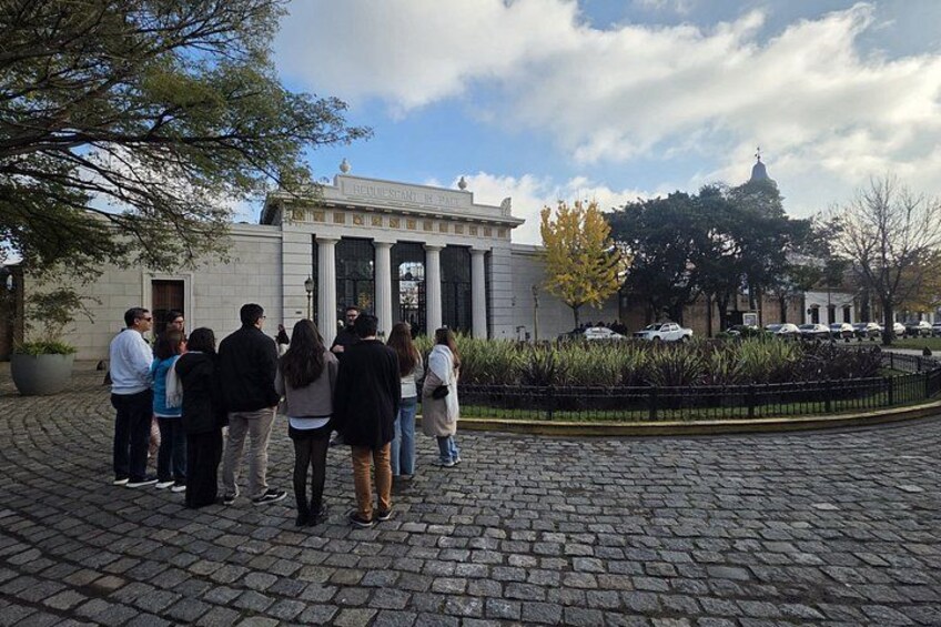 A group of people standing on a cobblestone path in front of a classical building with columns. 1