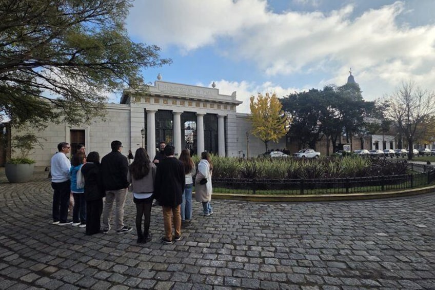 Un grupo de personas de pie sobre un camino adoquinado frente a un edificio clásico con columnas. 1