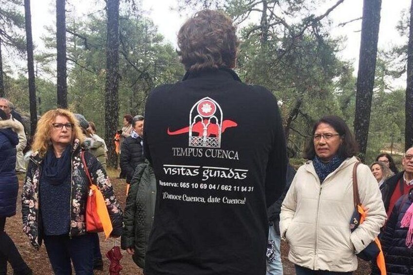 Un grupo de personas en un área boscosa, algunas con chaquetas, y una persona al fondo con una chaqueta negra que tiene un logotipo y texto. 5