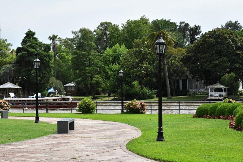A park with a paved walkway, green grass, a lamp post, a bench, and a boat on the water. 3