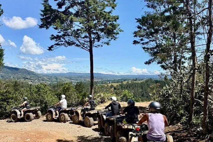 A group of people on all-terrain vehicles (ATVs) on a dirt path surrounded by trees, with a view of a mountainous landscape in the background. 1