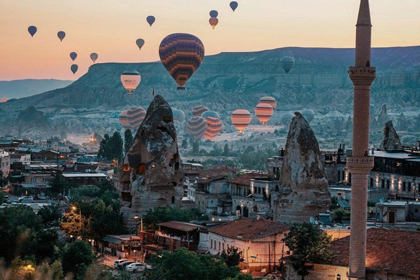 A cityscape with rock formations and a minaret, with hot air balloons in the sky. 1