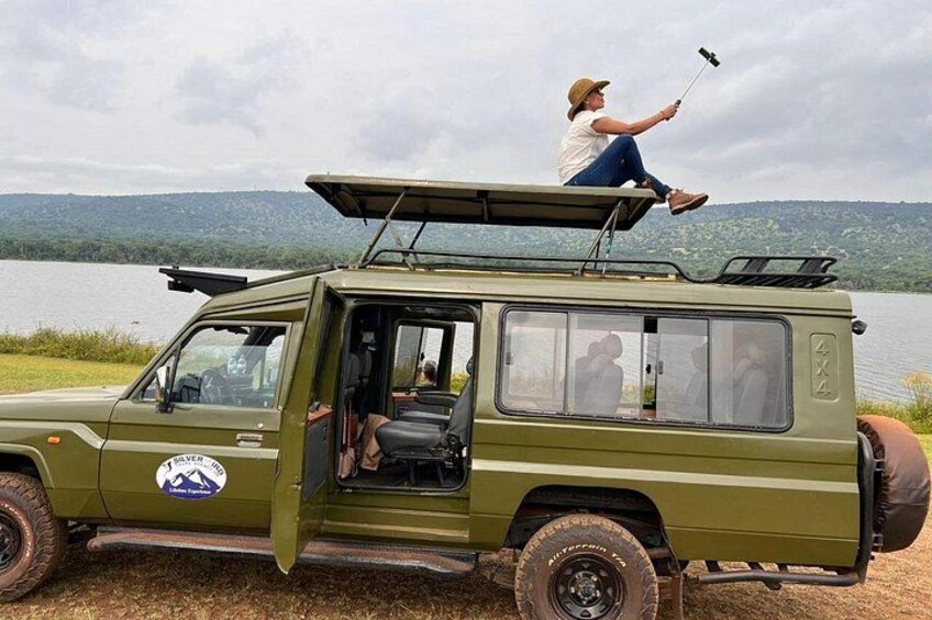 A green safari vehicle parked by a lake with a person taking a selfie. 3