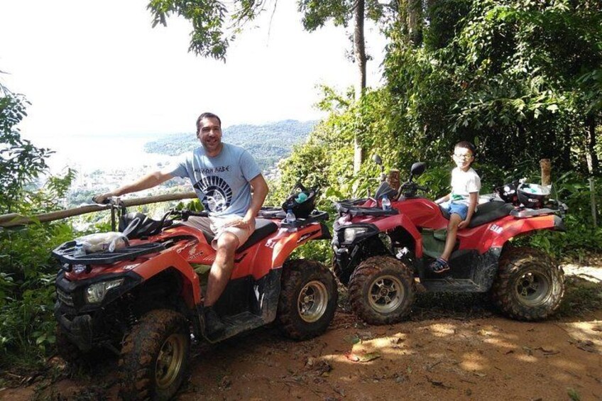 Deux personnes sur des quad rouges circulent sur un chemin de terre, avec en arrière-plan un plan d’eau et des collines. 3