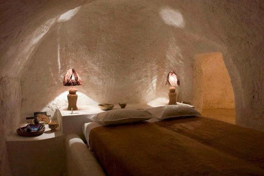 An interior of a cave-like structure with a bed, two lamps, and a small table with items. 1