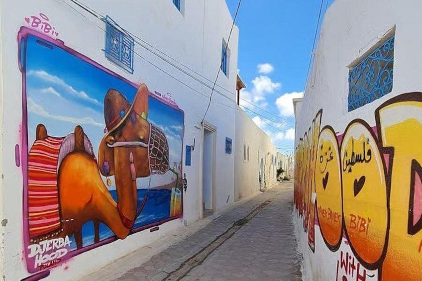 Une rue étroite bordée de bâtiments blancs, de graffitis colorés et d’une fresque représentant une femme et un âne. 3