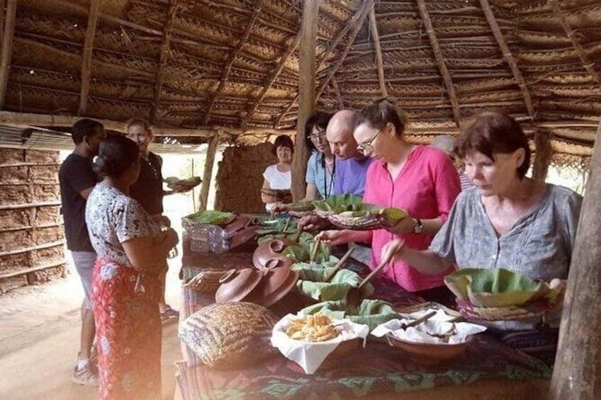 A group of people in a rustic setting with a thatched roof, engaged in a communal activity around a table with various food items. 5