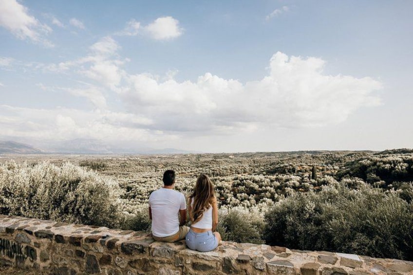 Two people sitting on a stone wall overlooking a desert landscape with sparse vegetation and distant mountains. 1