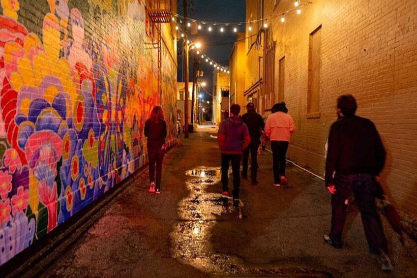 Une ruelle étroite, avec une fresque colorée sur le mur de gauche, un sol mouillé et des guirlandes lumineuses suspendues au-dessus. 2