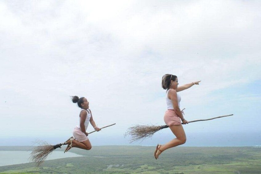 Two women jumping with brooms in mid-air against a backdrop of a cloudy sky and a distant landscape. 3