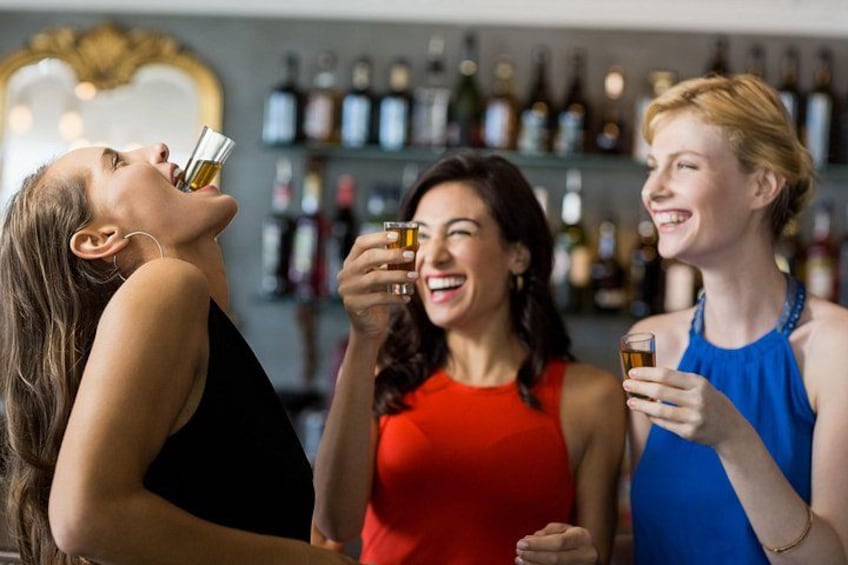 Tre donne che sorseggiano un drink in un bar, con scaffali pieni di bottiglie sullo sfondo. 4