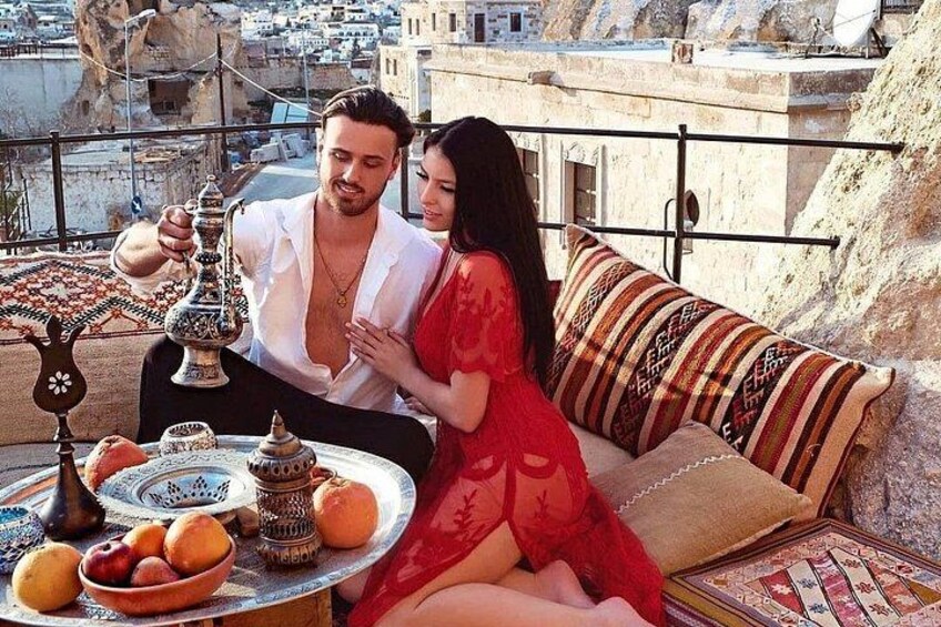 A couple sitting on a balcony with a table of traditional Middle Eastern food and drink, overlooking a cityscape. 1