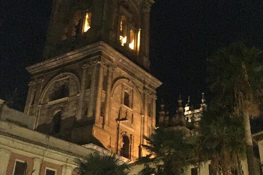 A historic tower with illuminated windows at night, surrounded by palm trees and buildings. 4