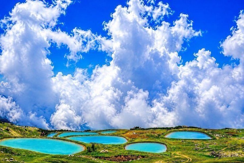 Eine Landschaft mit mehreren türkisfarbenen Seen, umgeben von grünen Hügeln und einem klaren, blauen Himmel mit wolkigen Wolken. 1